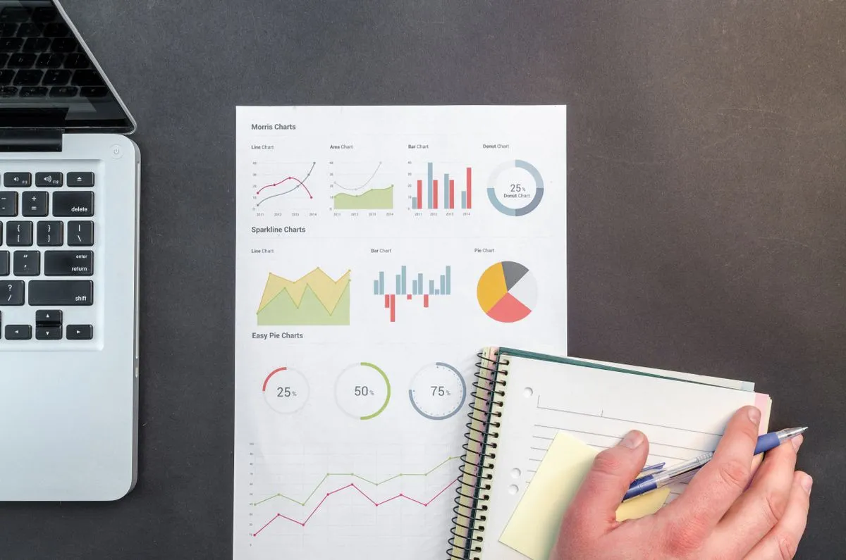 A notebook, a pen, and a laptop screen showing charts, tracking progress against a plan